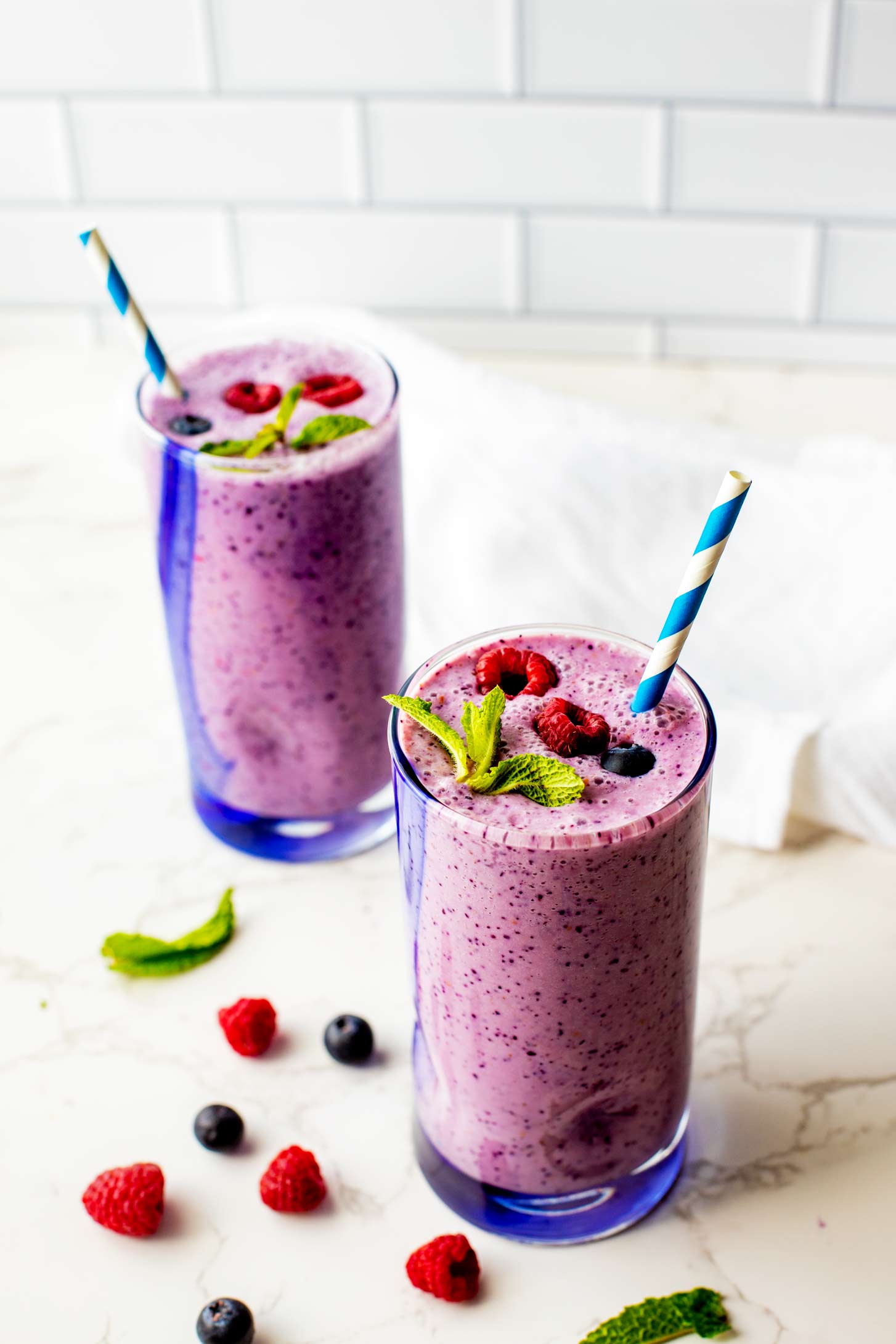 Photo of two glasses with a raspberry blueberry smoothie in them garnished with mint, raspberries, and blueberries.