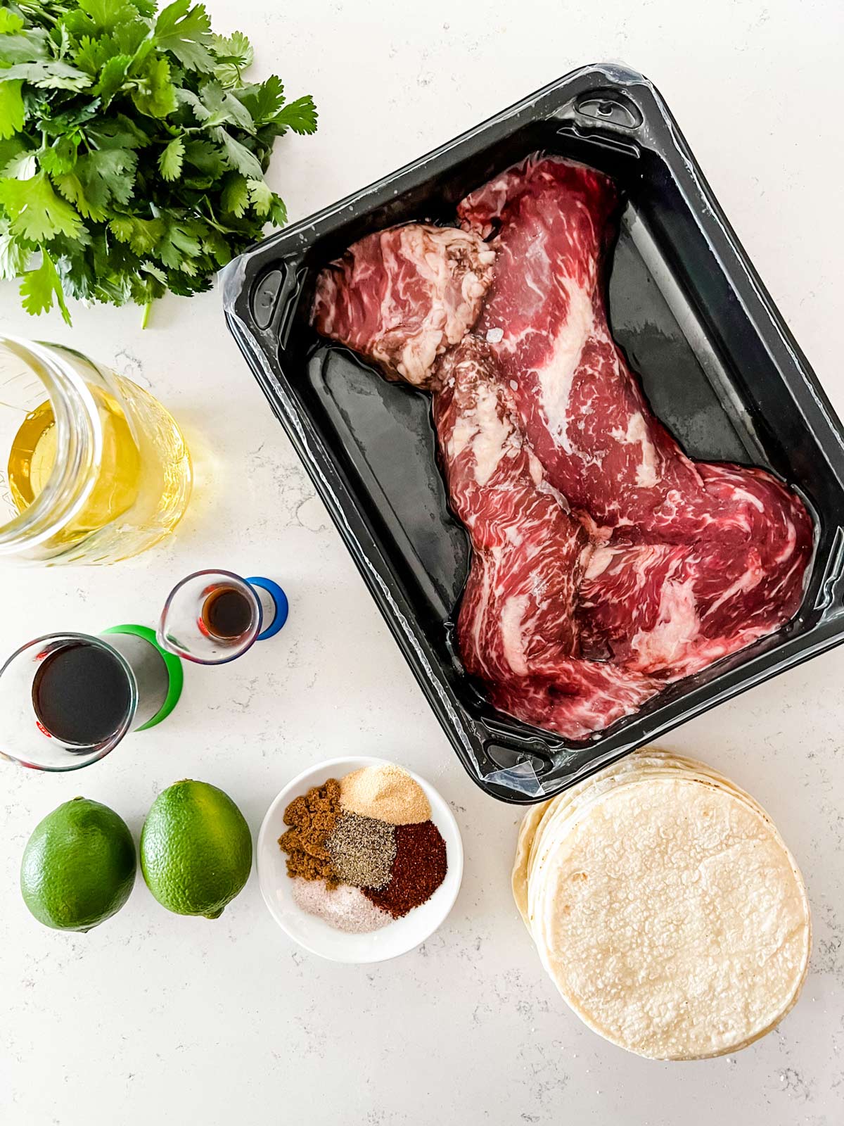 Overhead photo of hanger steak, tortillas, cilantro, oil, limes, seasonings, soy sauce and Worcestershire in prep containers.