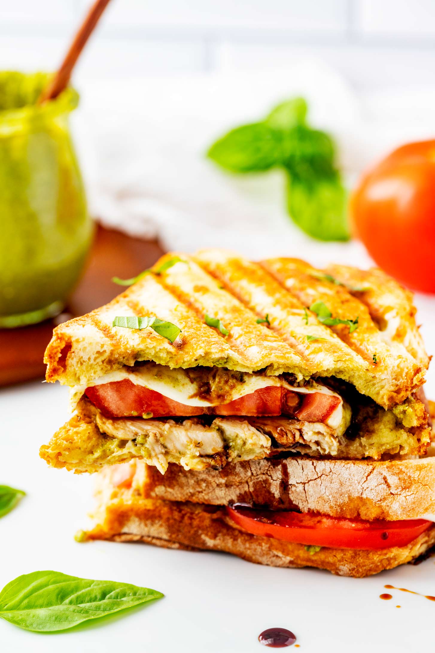 Side photo of a chicken caprese panini on a white surface with pesto, basil, and a tomato behind it.