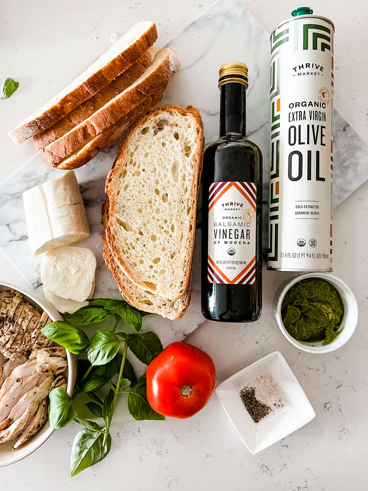 Overhead photo of olive oil, balsamic vinegar, pesto, bread, fresh mozzarella, cooked sliced chicken, tomato, basil, and seasonings.