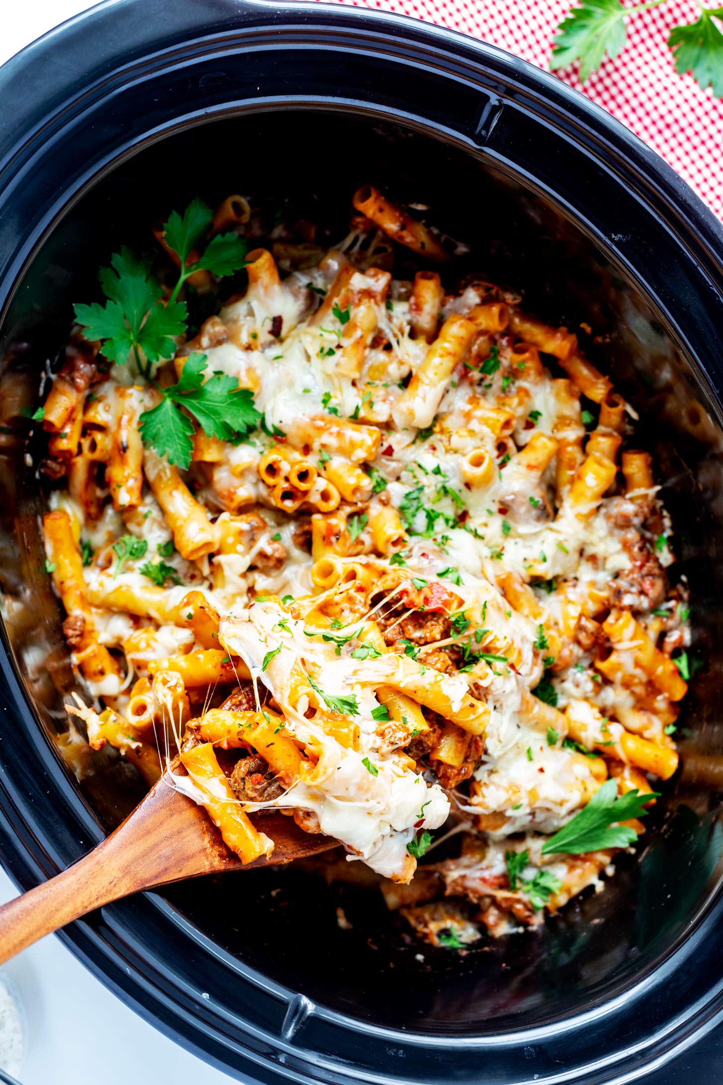 Overhead photo of crockpot baked ziti in a slow cooker with a wooden spoon pulling a cheesy serving from it.