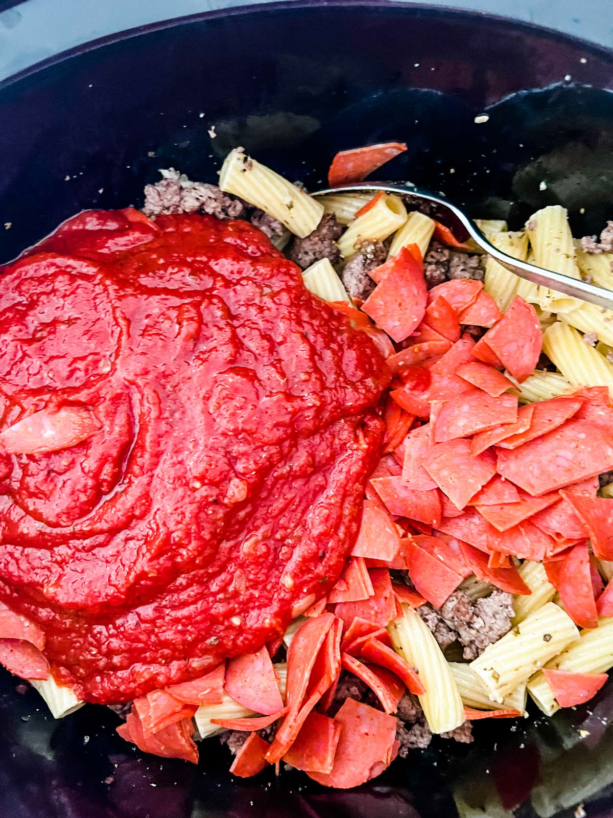 Marinara and chopped pepperoni being stirred into ground beef and pasta in a slow cooker.