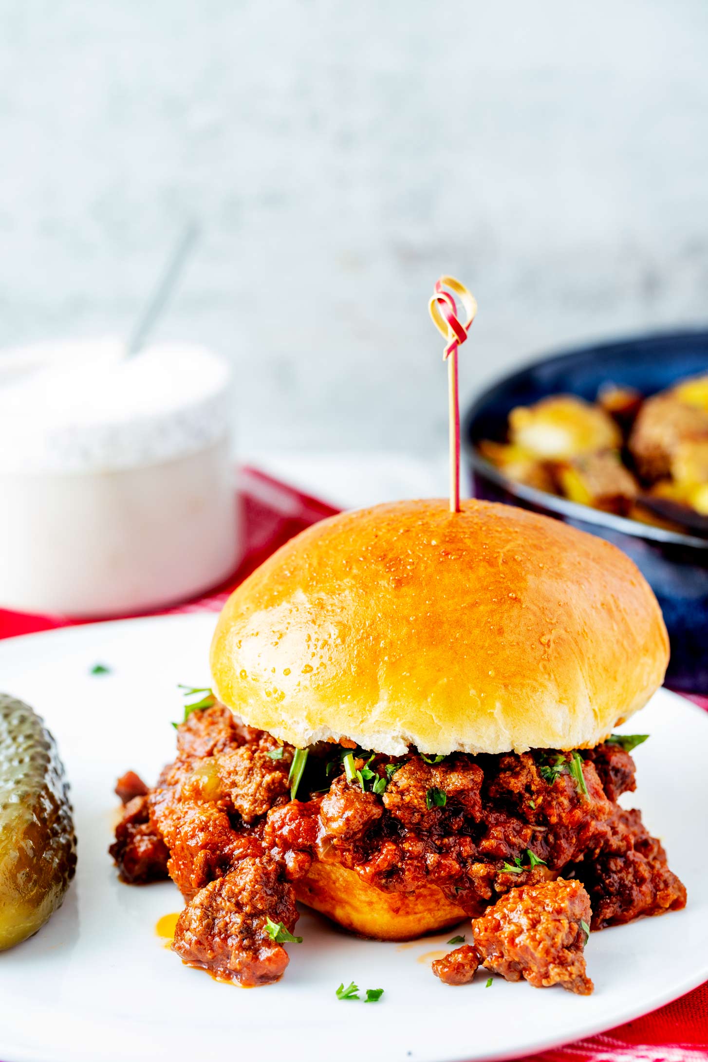 Side photo of slow cooker sloppy joes on a bun sitting on a white plate next to a pickle.