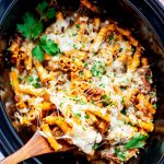 Square overhead photo of crockpot baked ziti in a slow cooker with a wooden spoon pulling a cheesy serving from it.