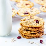 Square photo of stacked oatmeal craisin cookies sitting in front of a cooling rack of cookies next to a glass bottle of milk.