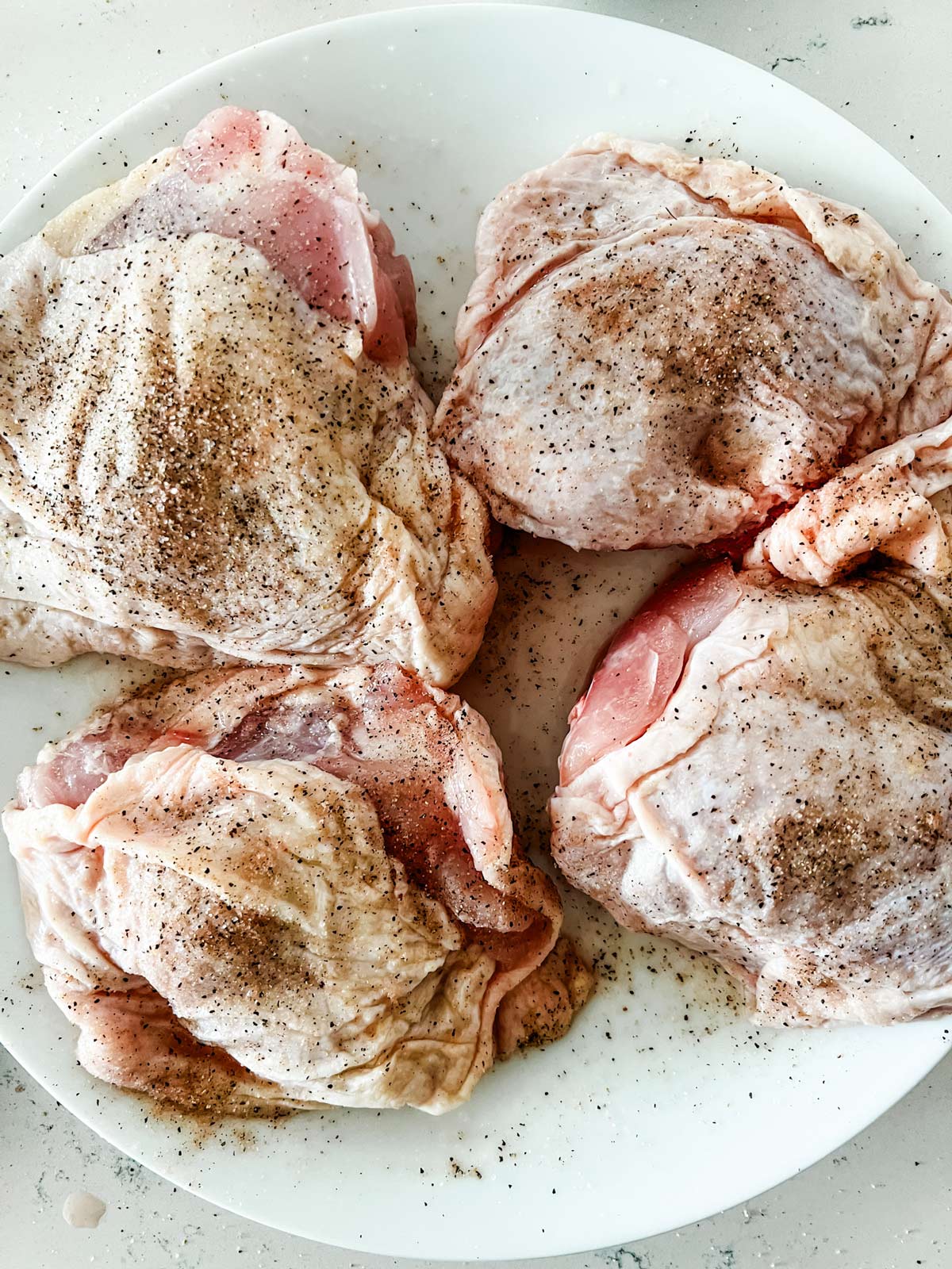 Bone-in chicken thighs seasoned with salt and pepper on a white plate.