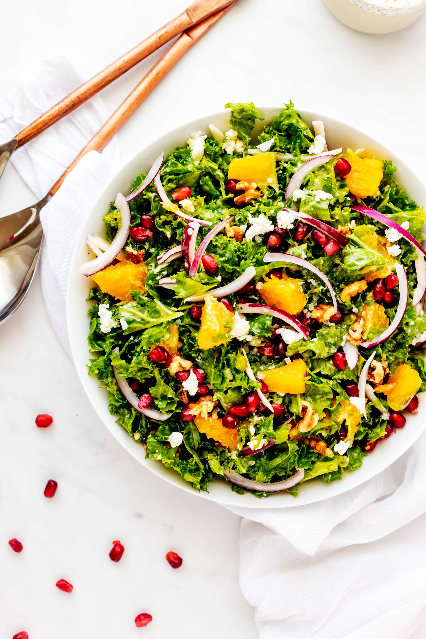 Overhead photo of a shallow bowl with kale pomegranate salad with salad serving spoon and pomegranate seeds sprinkled around.