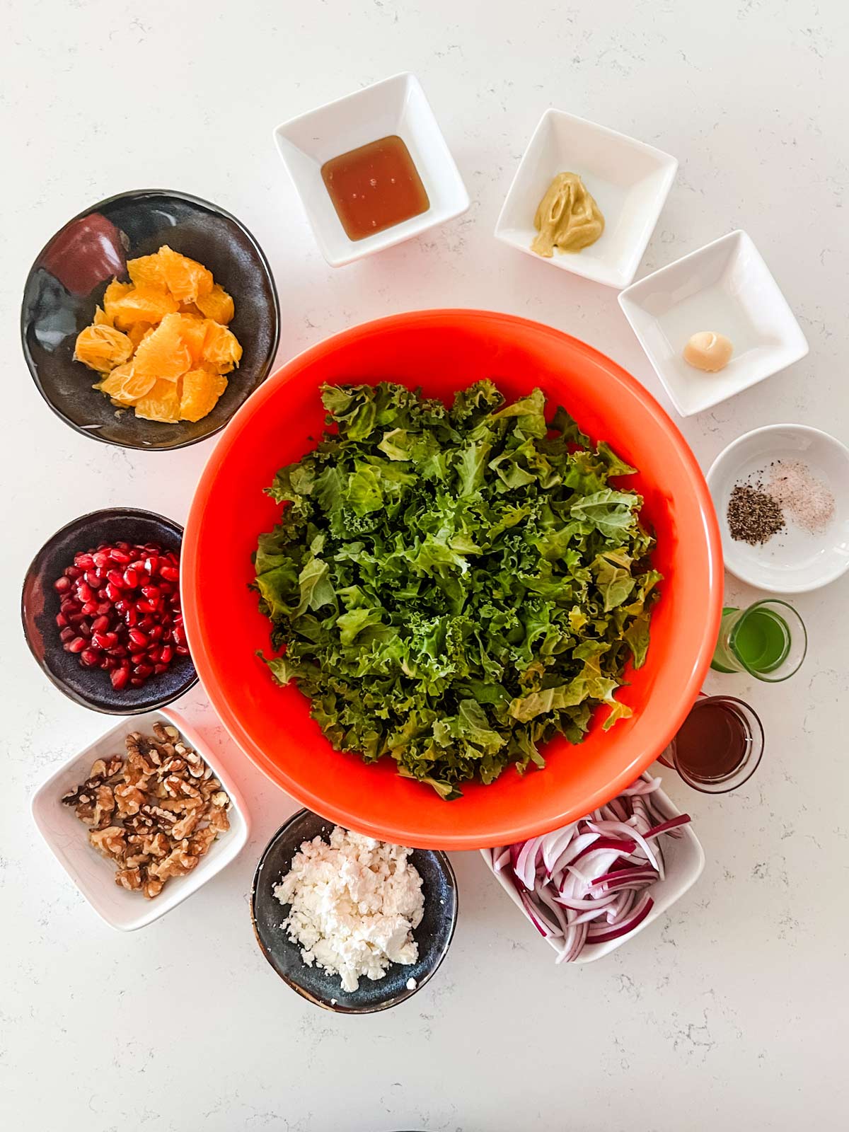 Kale, orange, pomegranate, walnuts, goat cheese, onion, and salad dressing ingredients in prep bowls.