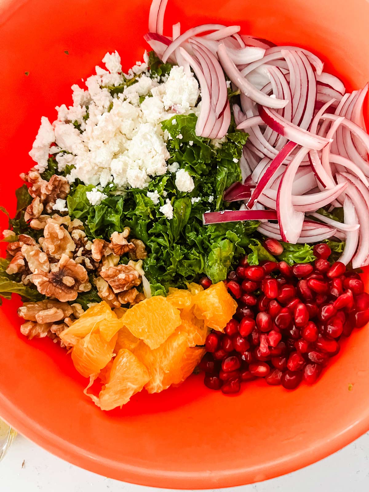 Kale in a bowl with pomegranate, orange, walnuts, goat cheese, and onion.