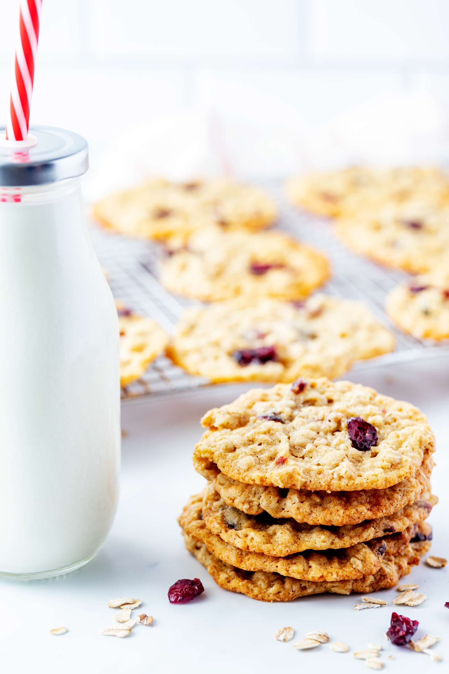 Photo of stacked oatmeal craisin cookies sitting in front of a cooling rack of cookies next to a glass bottle of milk.
