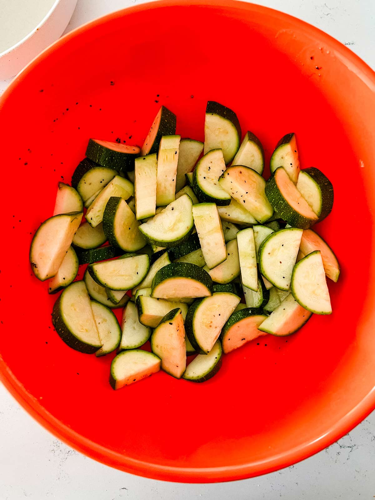 Overhead photo of halved and sliced zucchini tossed with seasonings and oil in an orange bowl.