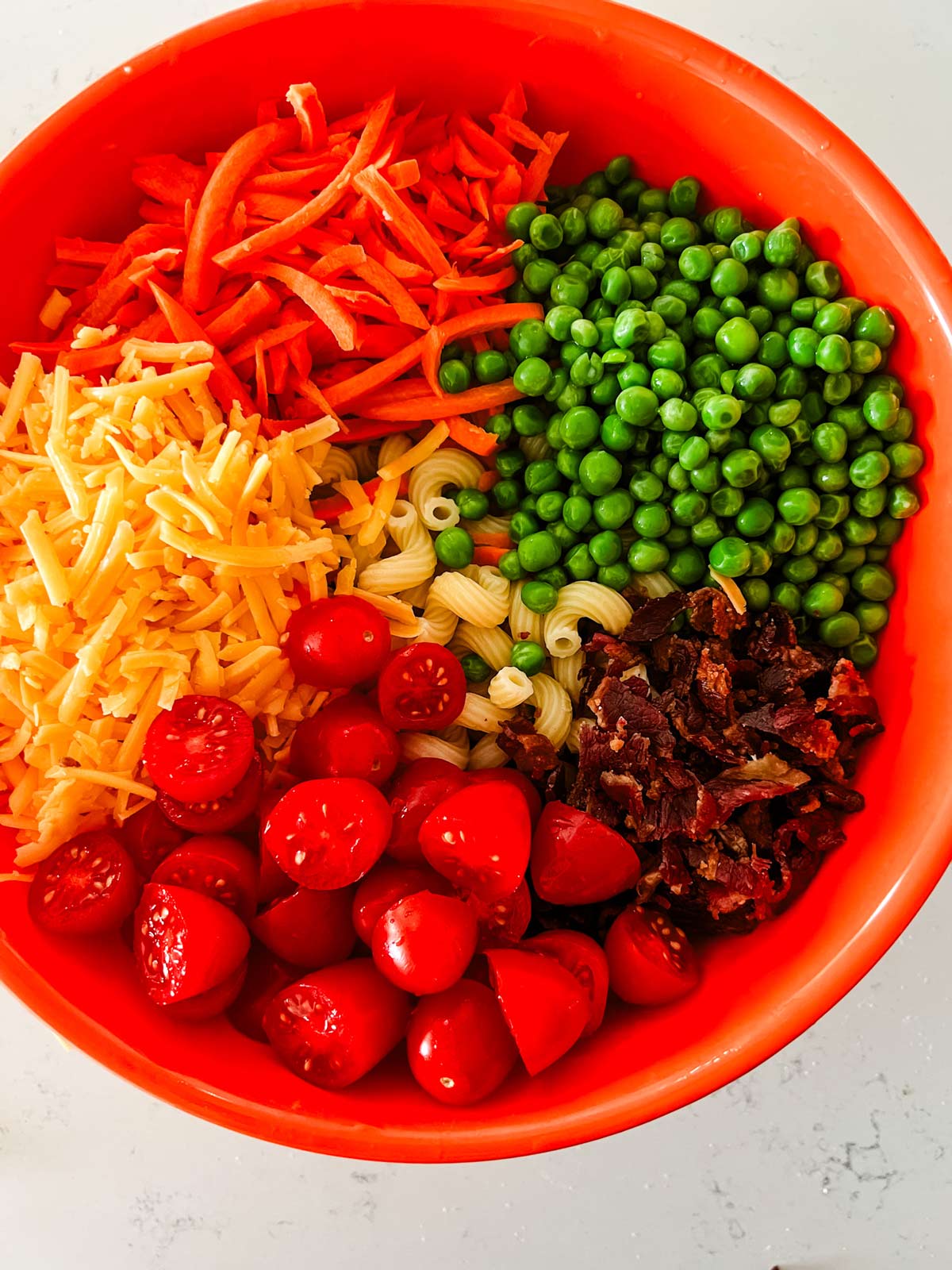 A bowl with pasta, bacon, cheese, tomatoes, carrots, and peas.