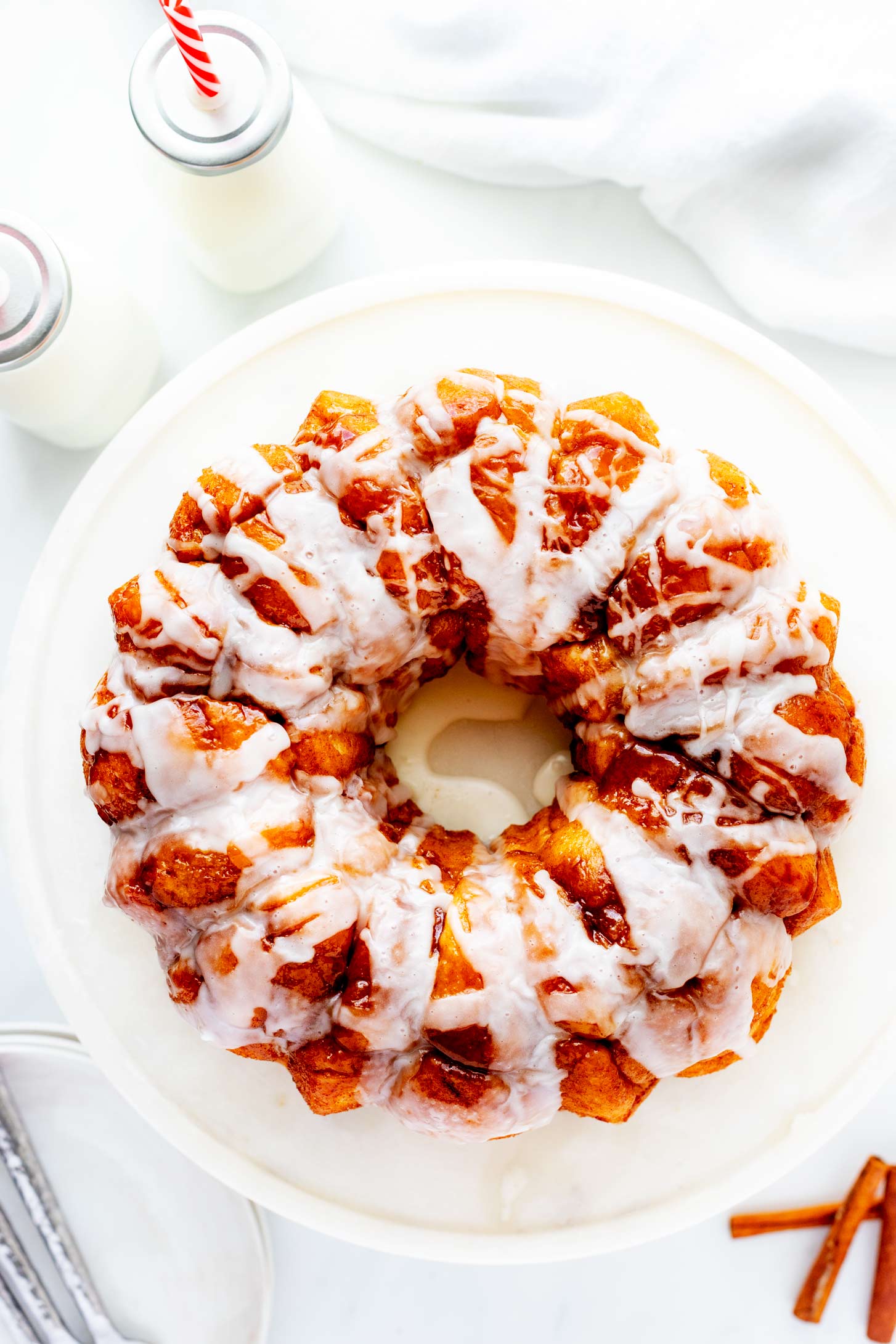 Overhead photo of bread machine monkey bread on a cake stand sitting next to two bottles of milks, serving plates, and cinnamon sticks.