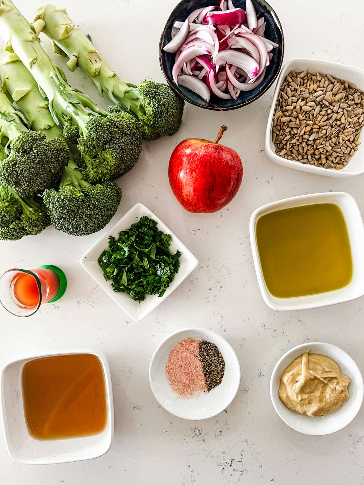 Overhead photo of broccoli, red onion, sunflower seeds, apple, parsley, dijon mustard, maple syrup, olive oil, cider vinegar, salt, and pepper.
