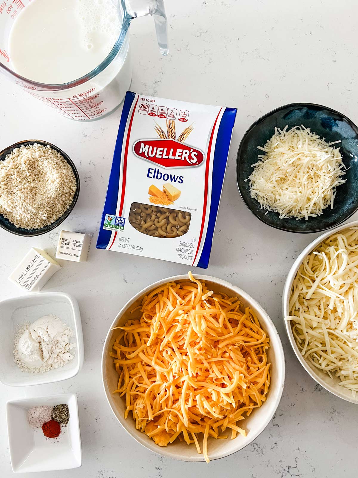 Overhead photo of three types of shredded cheese, milk, butter, flour, seasonings, elbow noodles, and breadcrumbs.