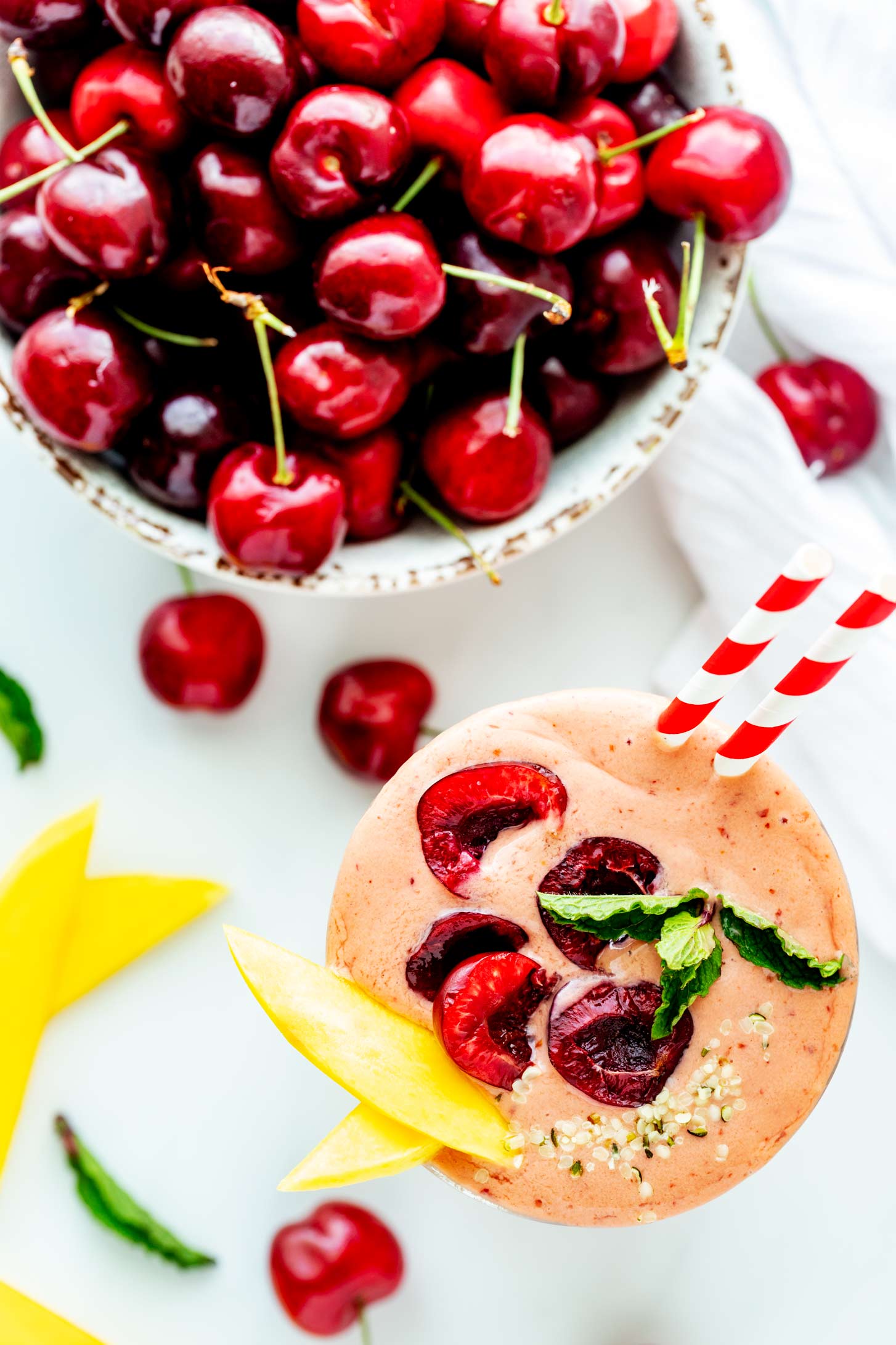Overhead photo of a cherry mango smoothie garnished with fresh cherries, mango slices, and mint and sitting next to a bowl of fresh cherries.