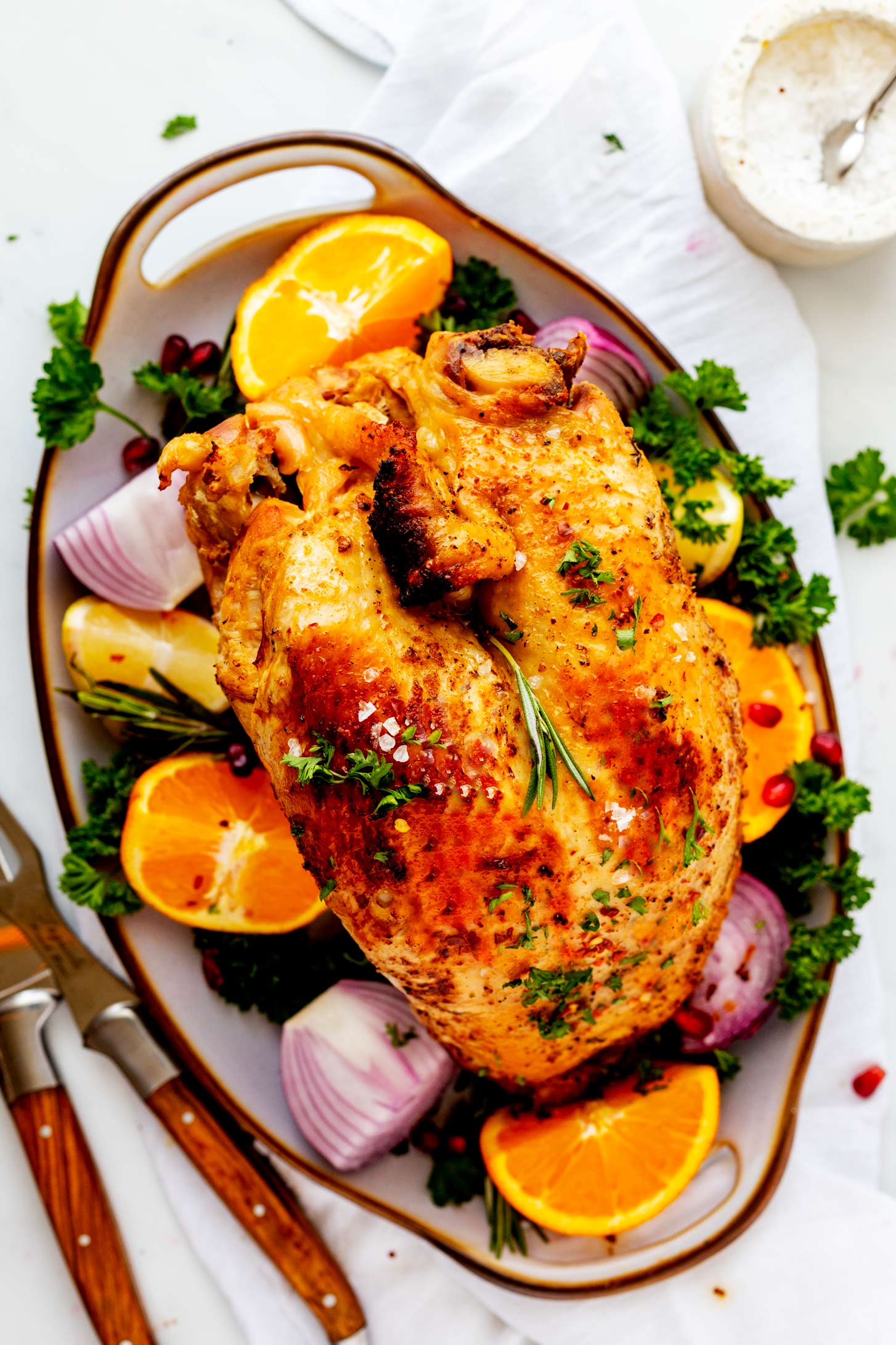 Overhead photo of a crockpot turkey breast on a serving platter surrounded by onion, parsley, lemon, and orange with carving knives next to it.