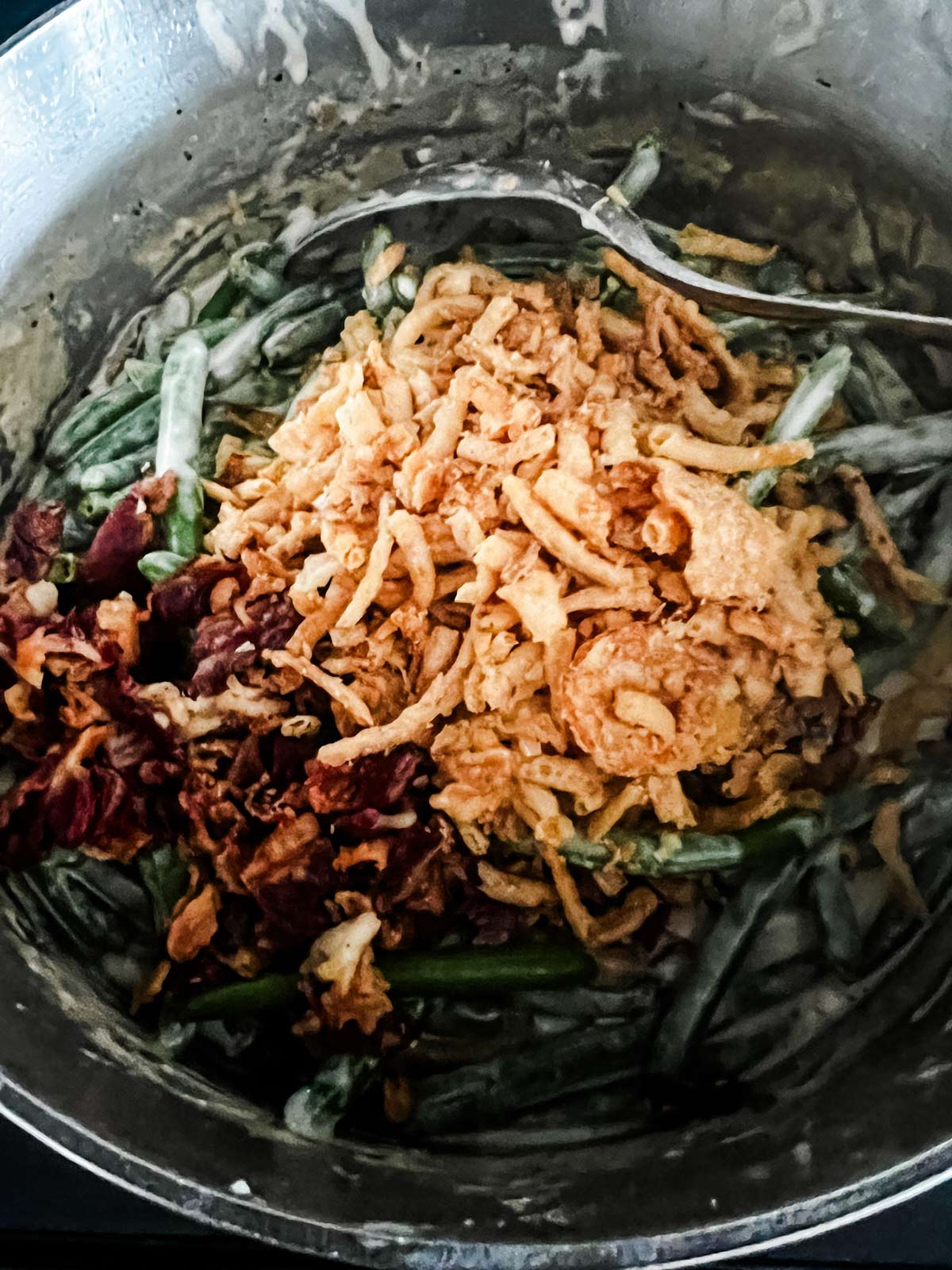 Green bean casserole with fried onions and bacon being stirred into it.