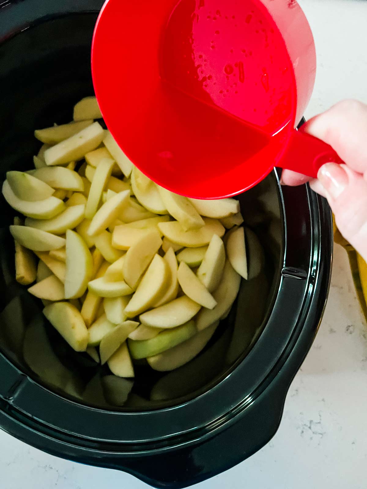 Lemon juice being poured on top of sliced apples in a slow cooker.