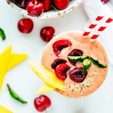 Overhead square photo of a cherry mango smoothie garnished with fresh cherries, mango slices, and mint and sitting next to a bowl of fresh cherries.