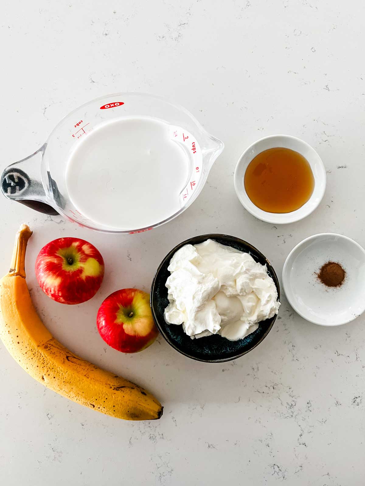 Overhead photo of oat milk, yogurt, apple, banana, honey, and cinnamon.