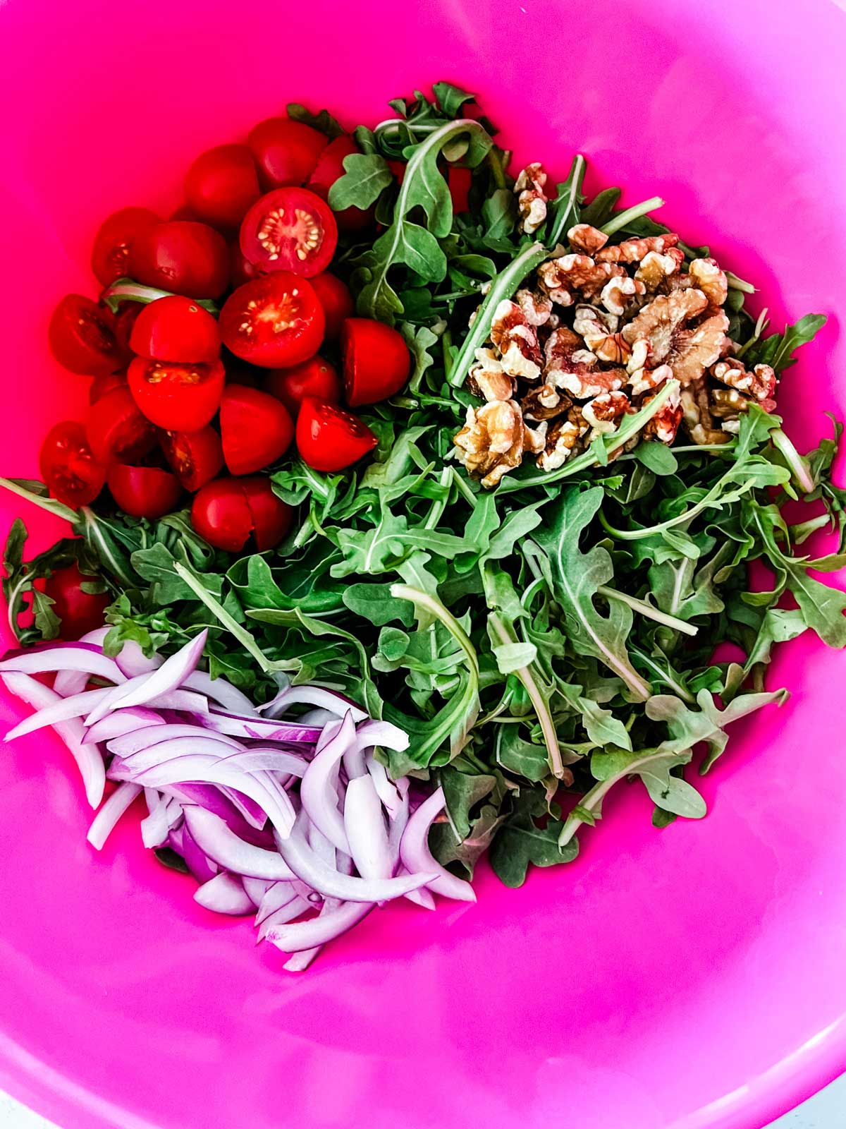 Arugula, tomato, walnuts, and red onion in a pink bowl.