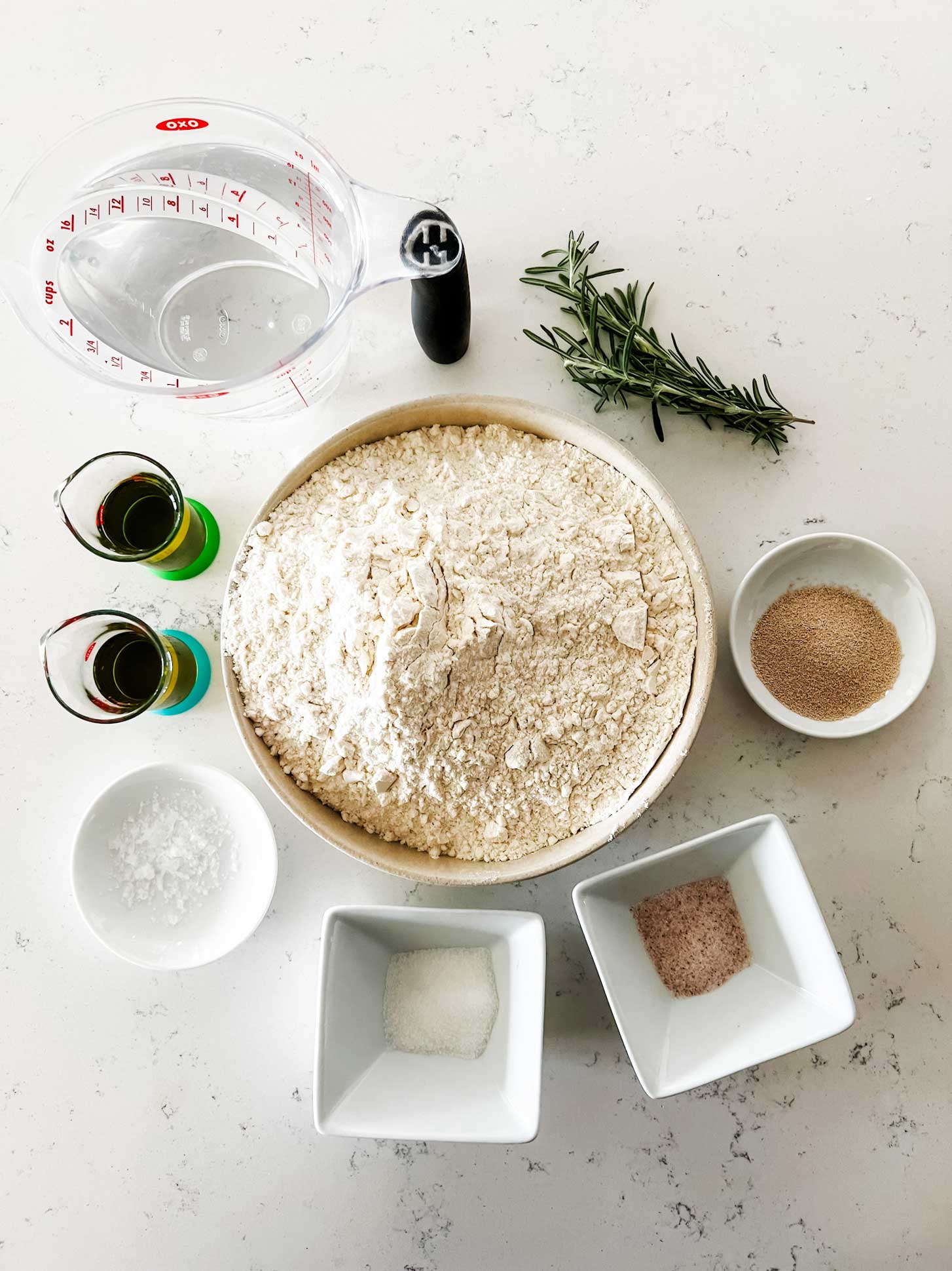 Overhead photo of flour, water, yeast, salt, sugar, flakey salt, oil, and rosemary.