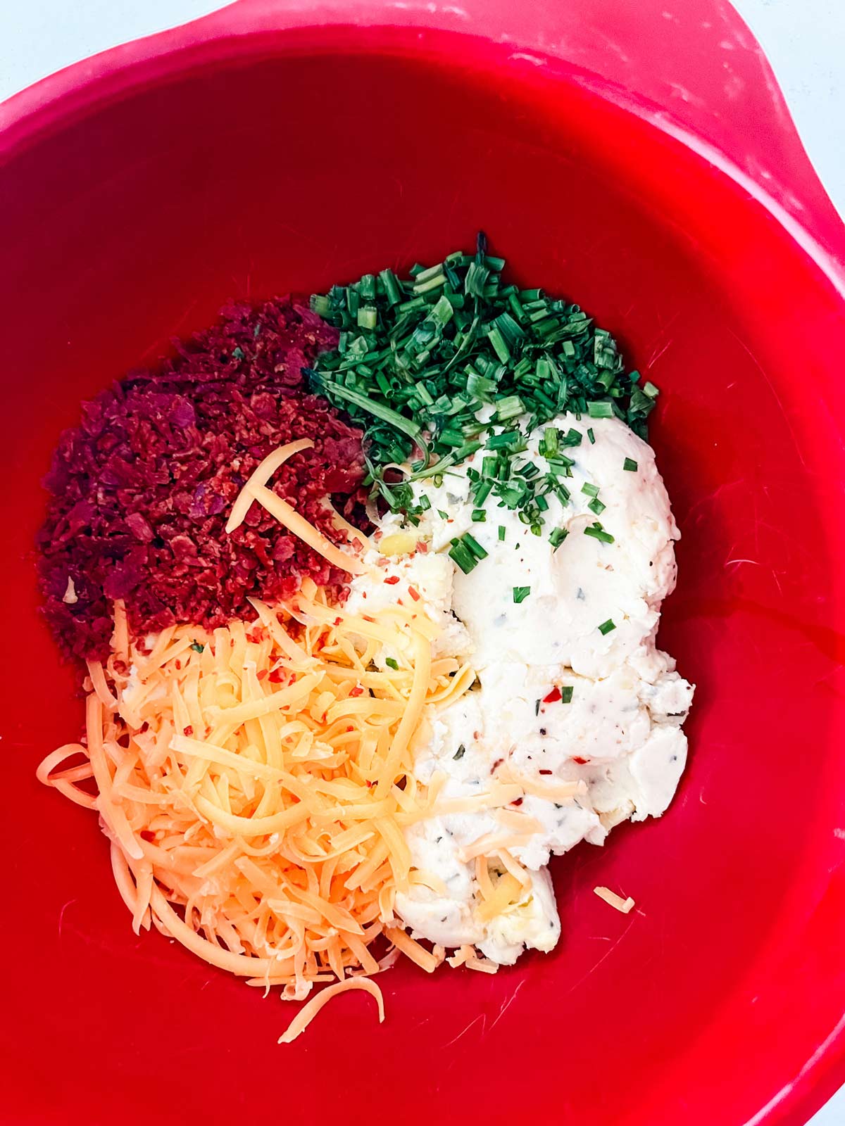 Mashed potatoes, bacaon, chives, and shredded cheese in a red bowl.