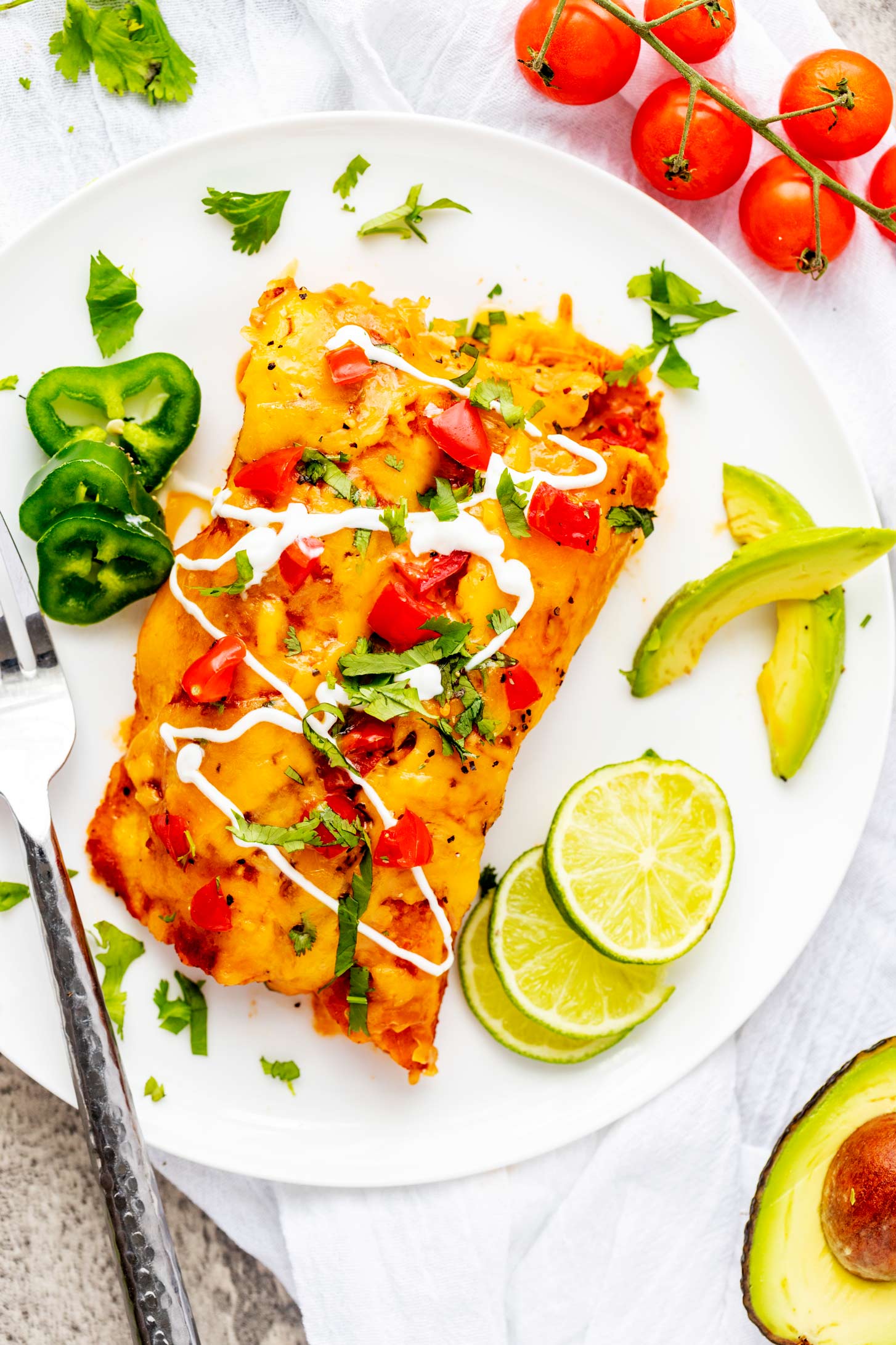 Overhead photo of slow cooker chicken enchiladas on a white place garnished with cilantro, lime, tomato and avocado.