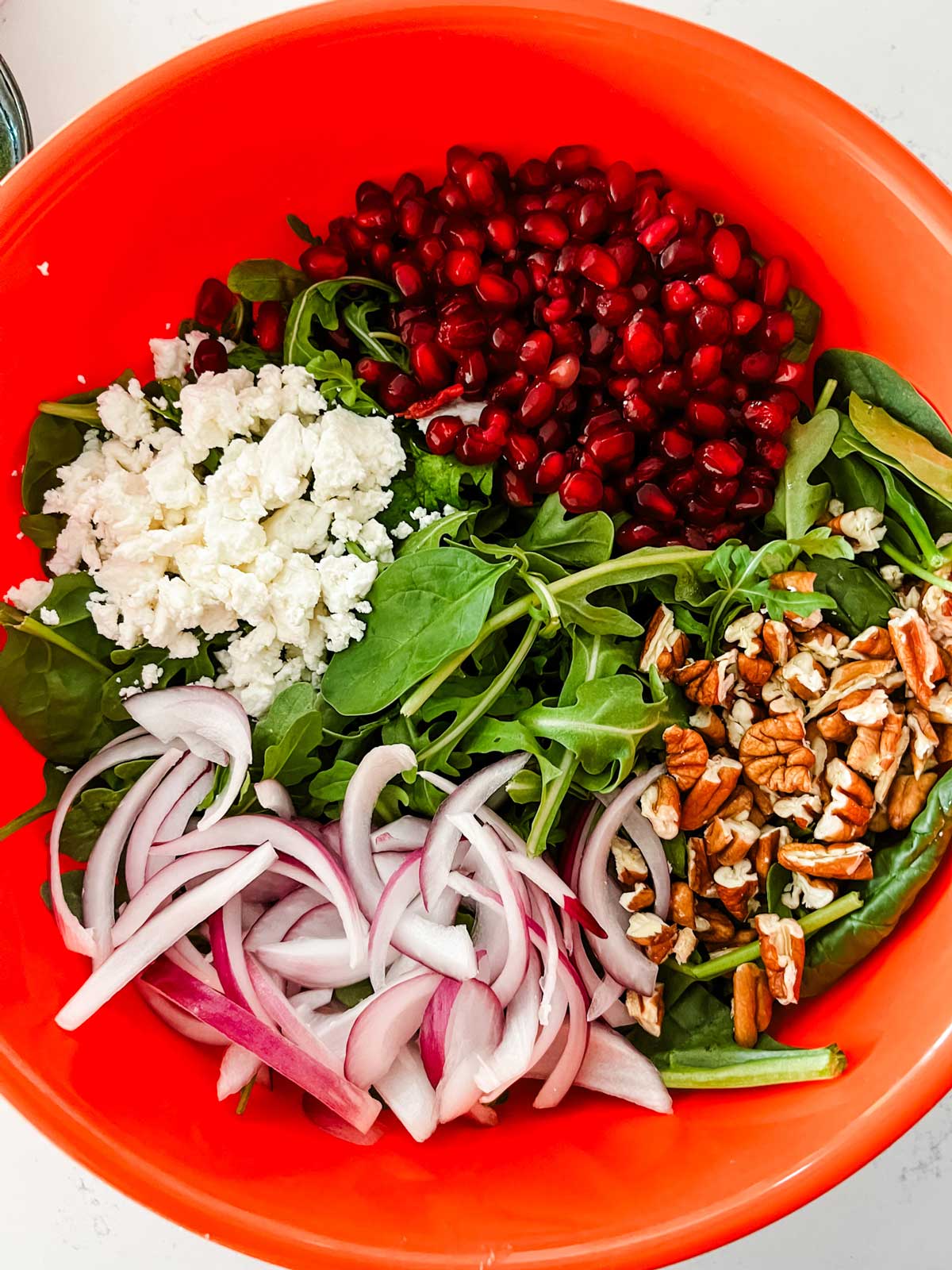 Overhead photo of spinach, arugula, pomegranate, goat cheese, and pecans in an orange bowl.