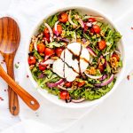 Square photo of an arugula burrata salad in a large bowl with salad serving spoons next to it.
