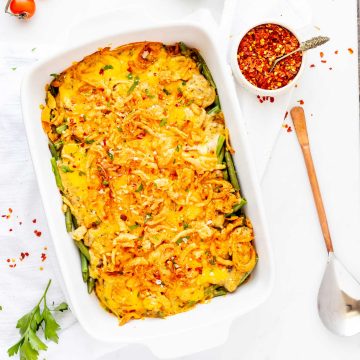 Overhead photo of cheesy green bean casserole in a white casserole dish next to a serving spoon and small dish of red pepper flakes.