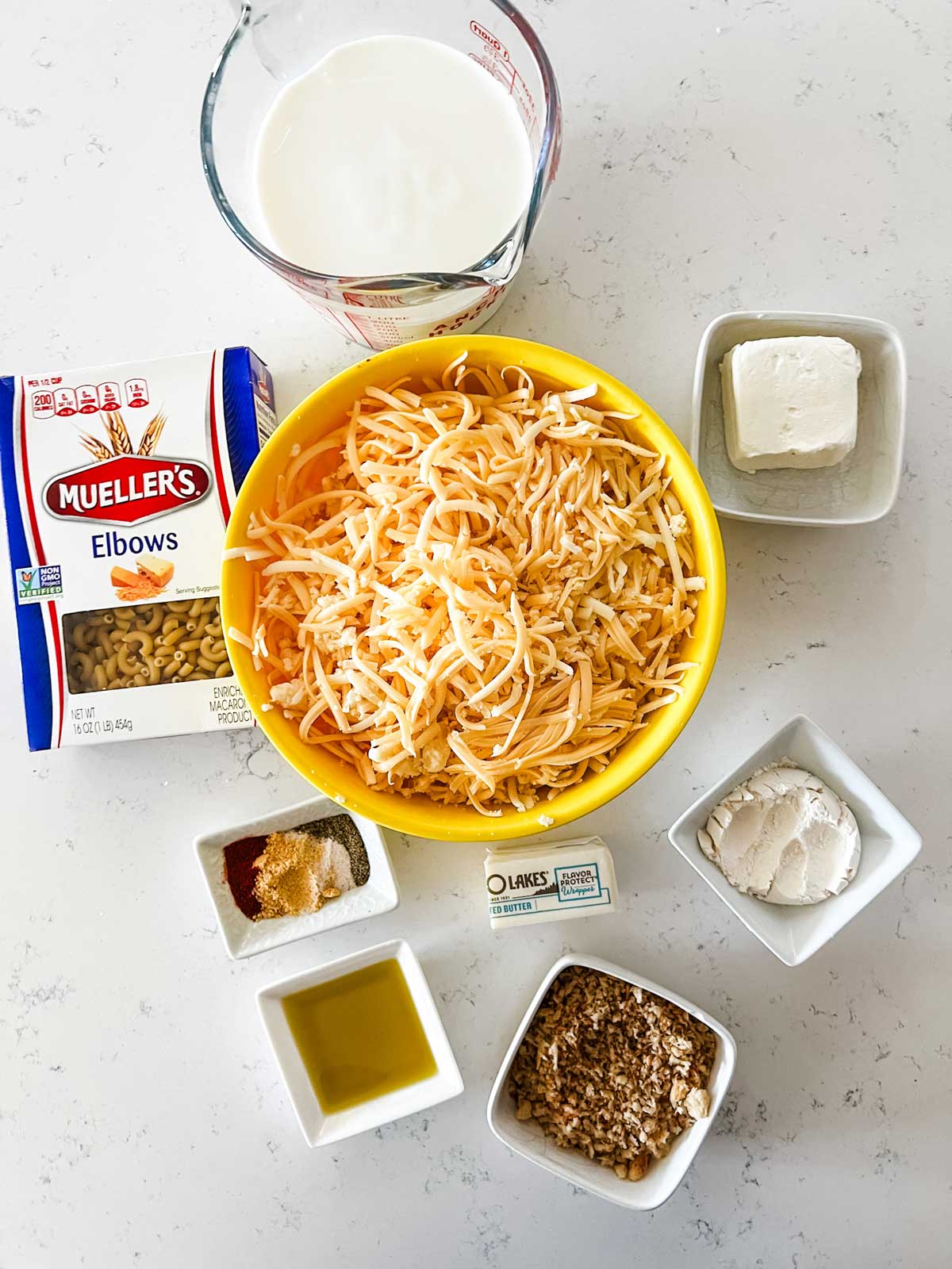 Overhead photo of milk, elbow noodles, white cheddar, smoked gouda, cream cheese, breadcrumbs, seasonings, oil, flour, and butter.