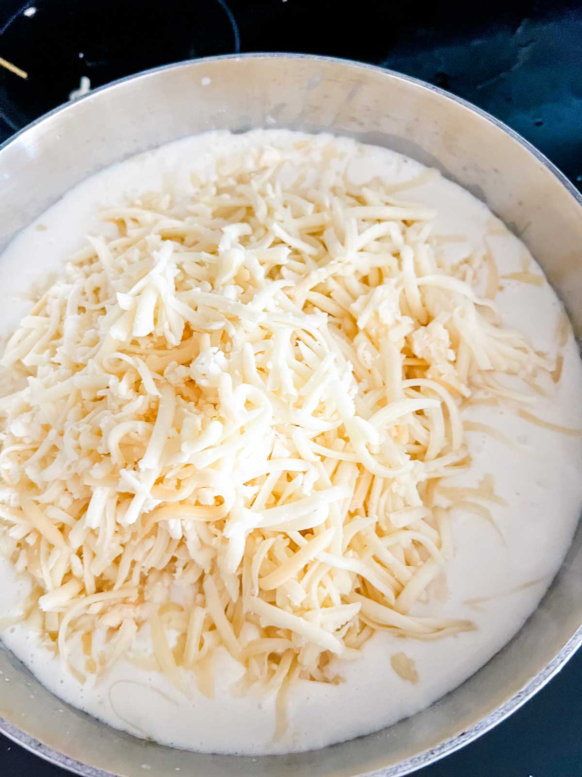 Shredded cheese added to a cream sauce in a saucepan.