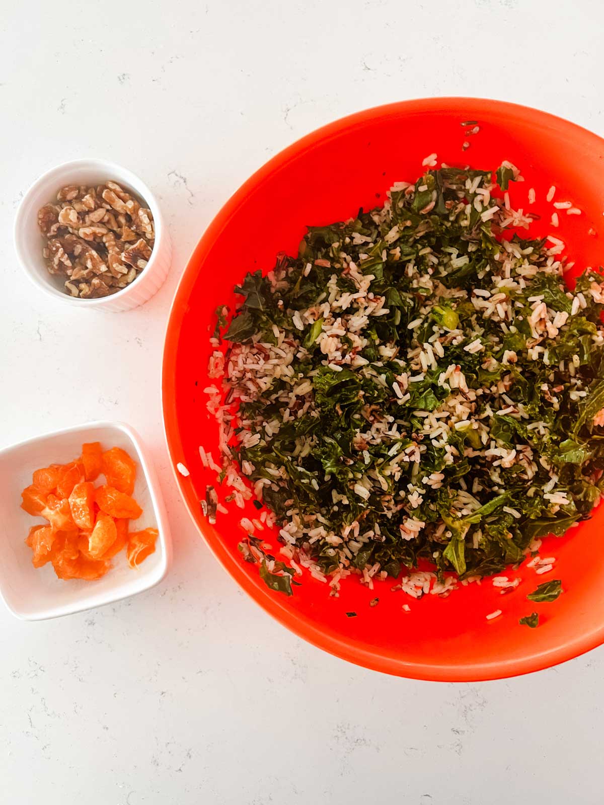 Overhead photo of wild rice and kale in an orange bowl with oranges and pecans in small prep bowls next to it.