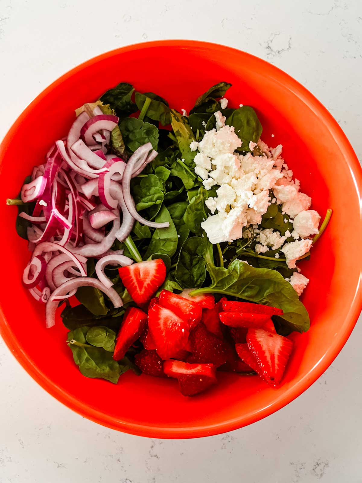 A large orange bowl with baby greens, goat cheese, red onion, and strawberries.