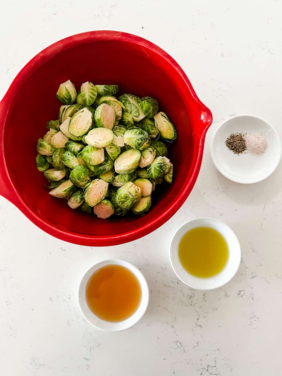 Overhead photo of brussels sprouts in a red bowl with small dishes of maple syrup, oil, and seasonings next to it.