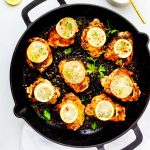 Square photo of cast iron chicken thighs in a large skillet garnished with lemon slices and parsley.