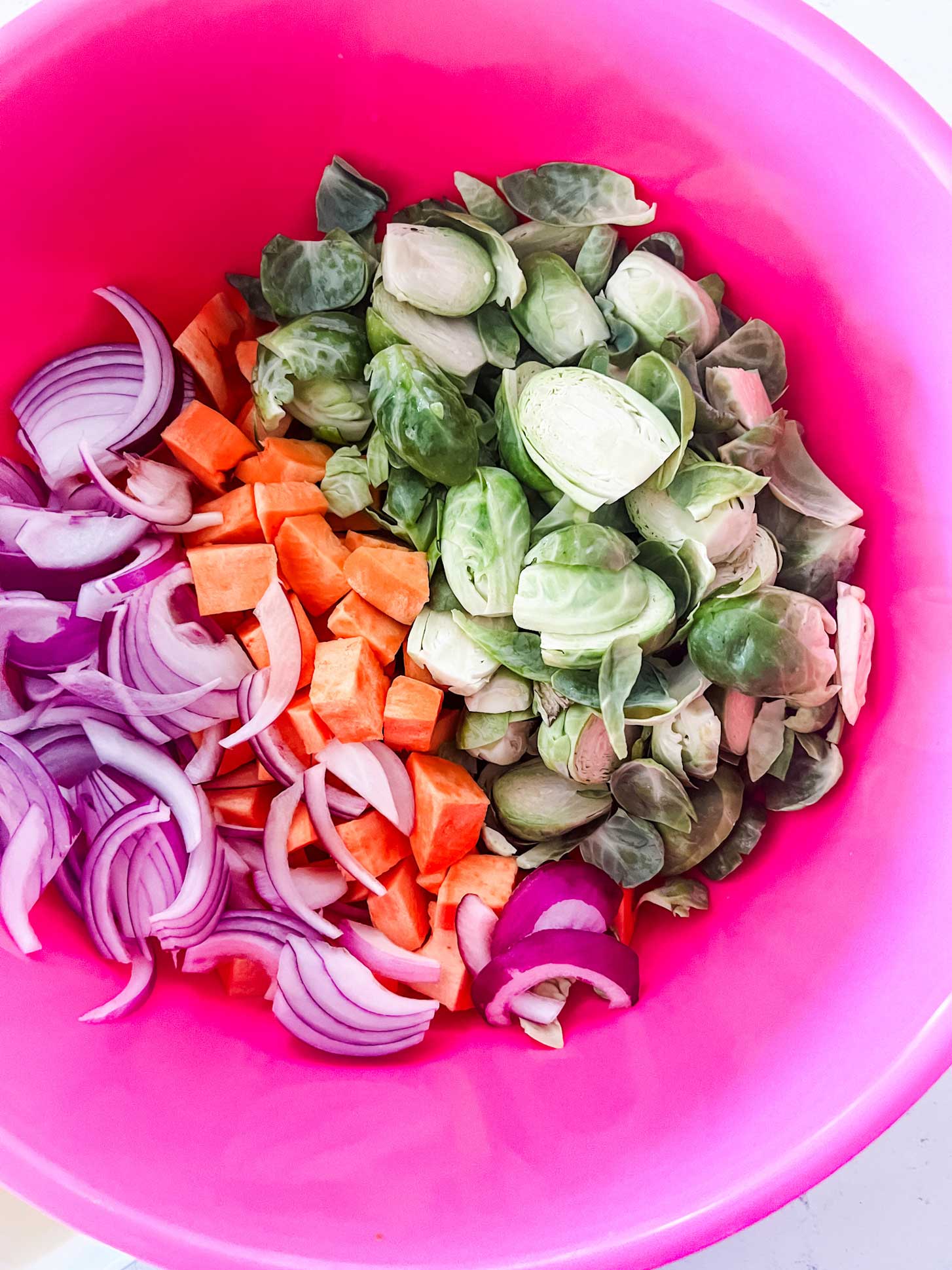 A pink bowl with Brussels sprouts, sweet potatoes, and red onion.