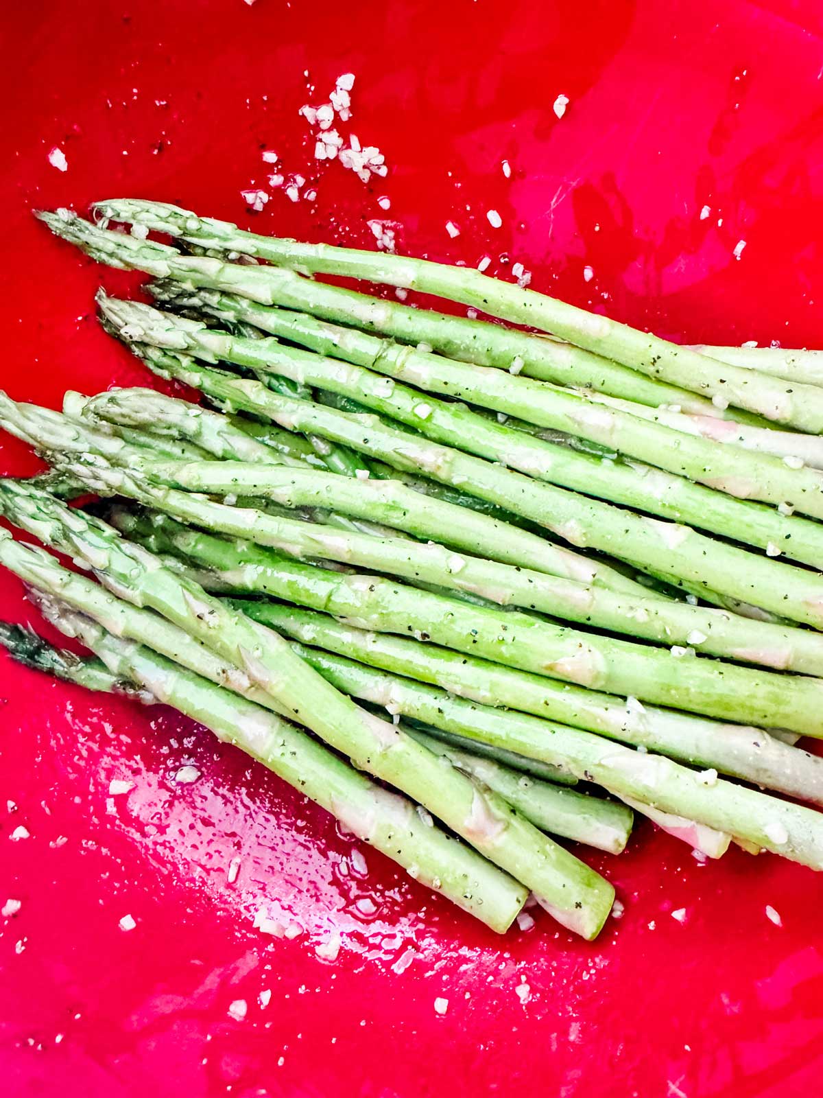 Asparagus in a red bowl that has been tossed with garlic, oil, and seasonings.