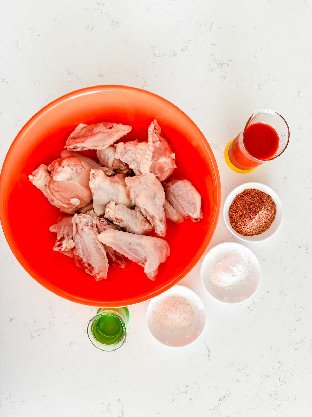 Overhead photo of chicken wings in an orange bowl surrounded by oil, seasonings, and buffalo sauce.