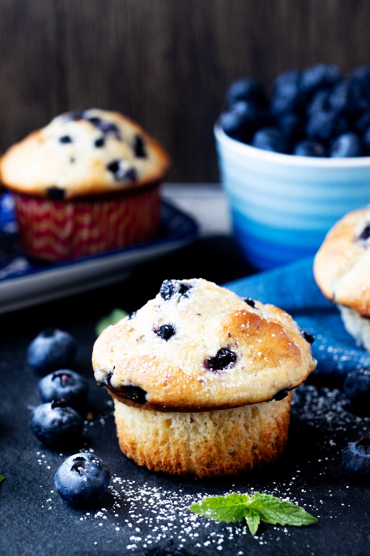 Side photo of buttermilk blueberry muffins sitting with a bowl of fresh blueberries.