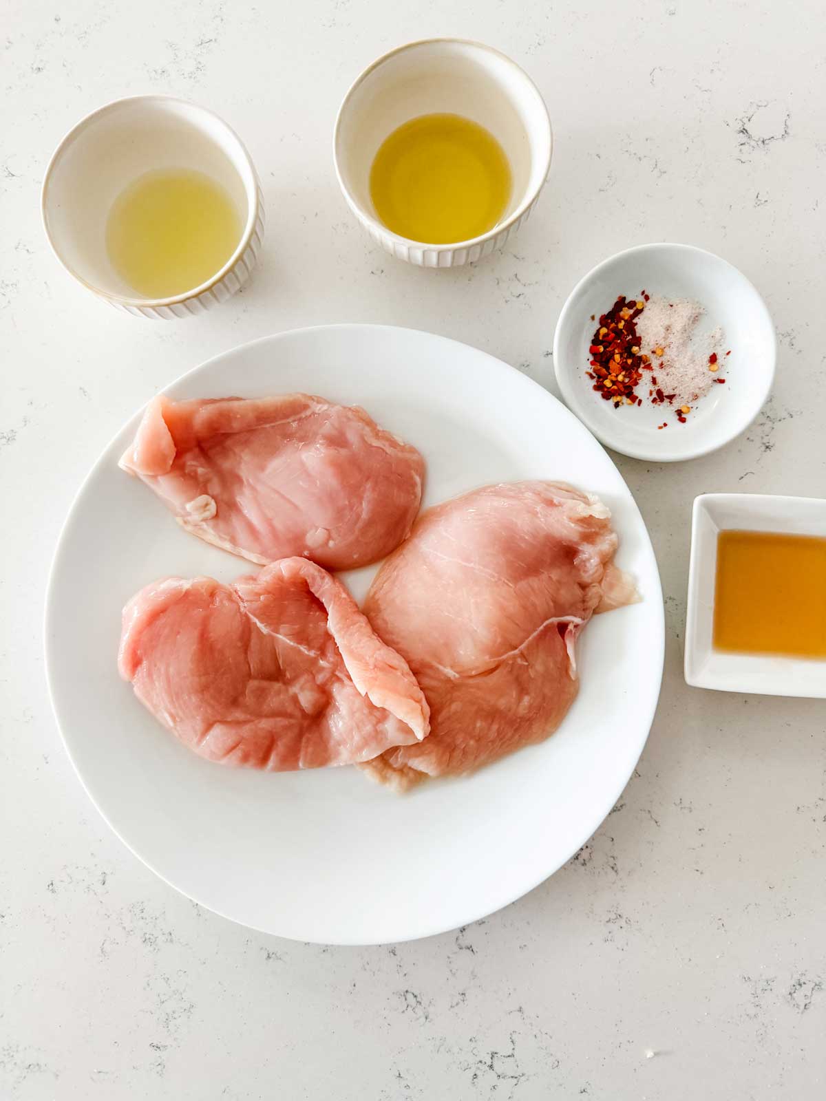 A plate of boneless chicken breasts sitting next to honey, seasonings, lime juice, and oil.