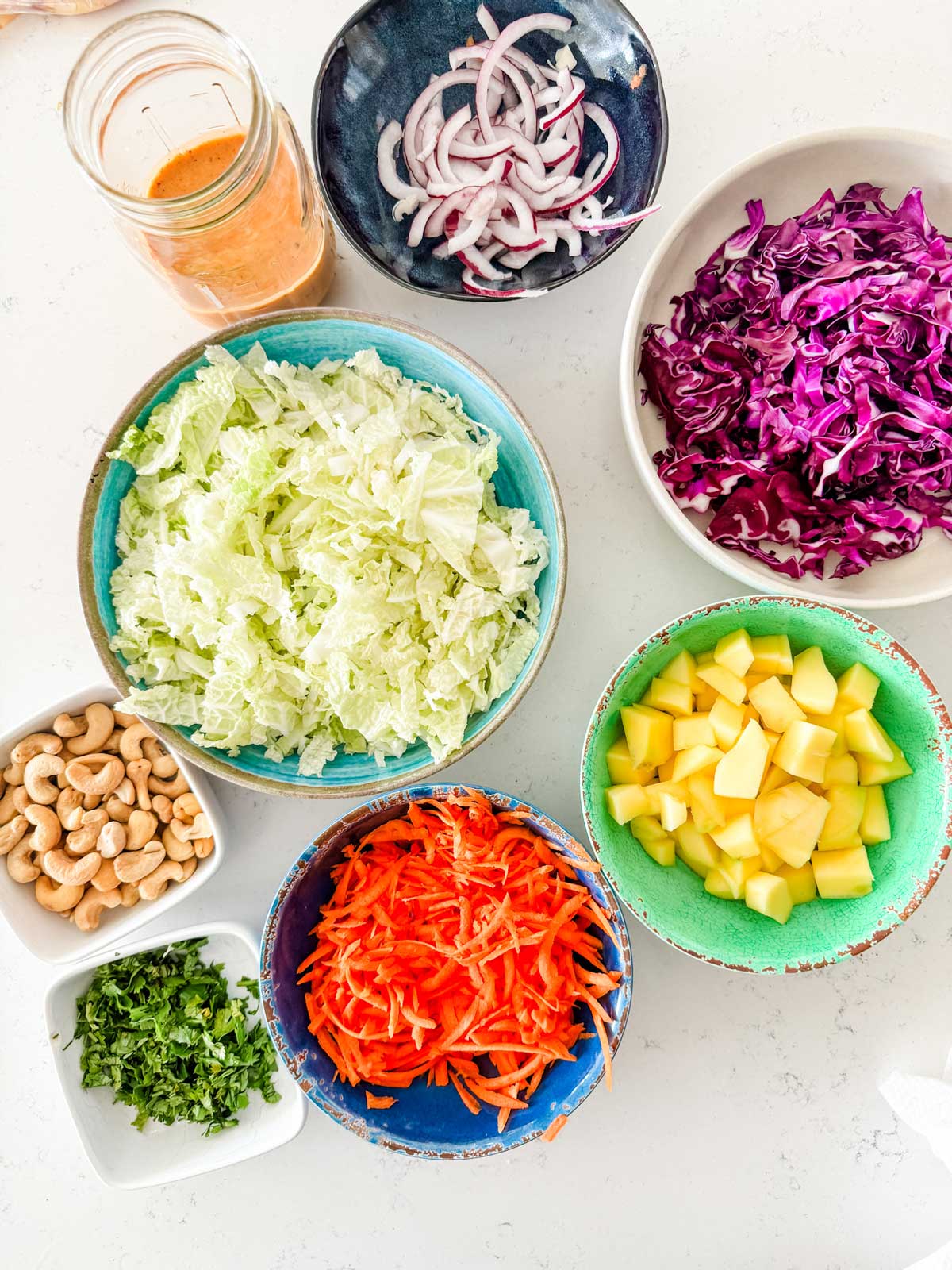 Overhead photo of salad dressing, napa cabbage, red onion, cabbage, mango, shredded carrots, cilantro and cashews.
