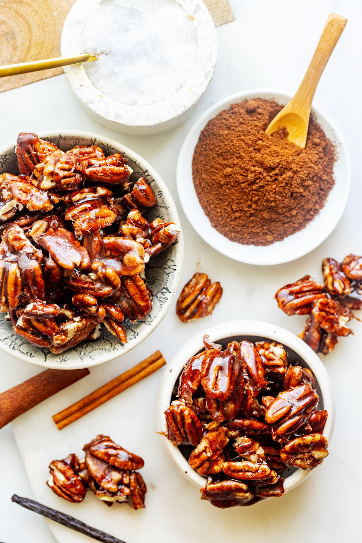 Overhead photo of two bowls of candied spiced pecans next to spices, salt, and scattered pecans.