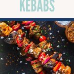 Grilled Blackstone kebabs with seasoned meat, red onion, and bell peppers on skewers, garnished with herbs and salt flakes. Served beside bowls of coarse salt and crushed red pepper. Text above reads Blackstone Kebabs.