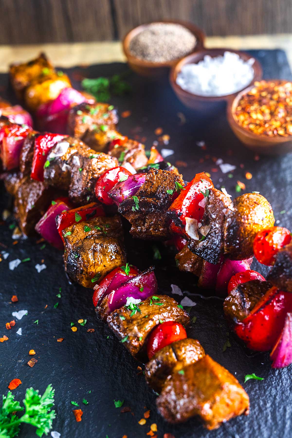 Two skewers of grilled beef, red onion, and red bell pepper on a dark slate surface, garnished with chopped herbs. Small bowls of salt, pepper, and chili flakes are in the background.