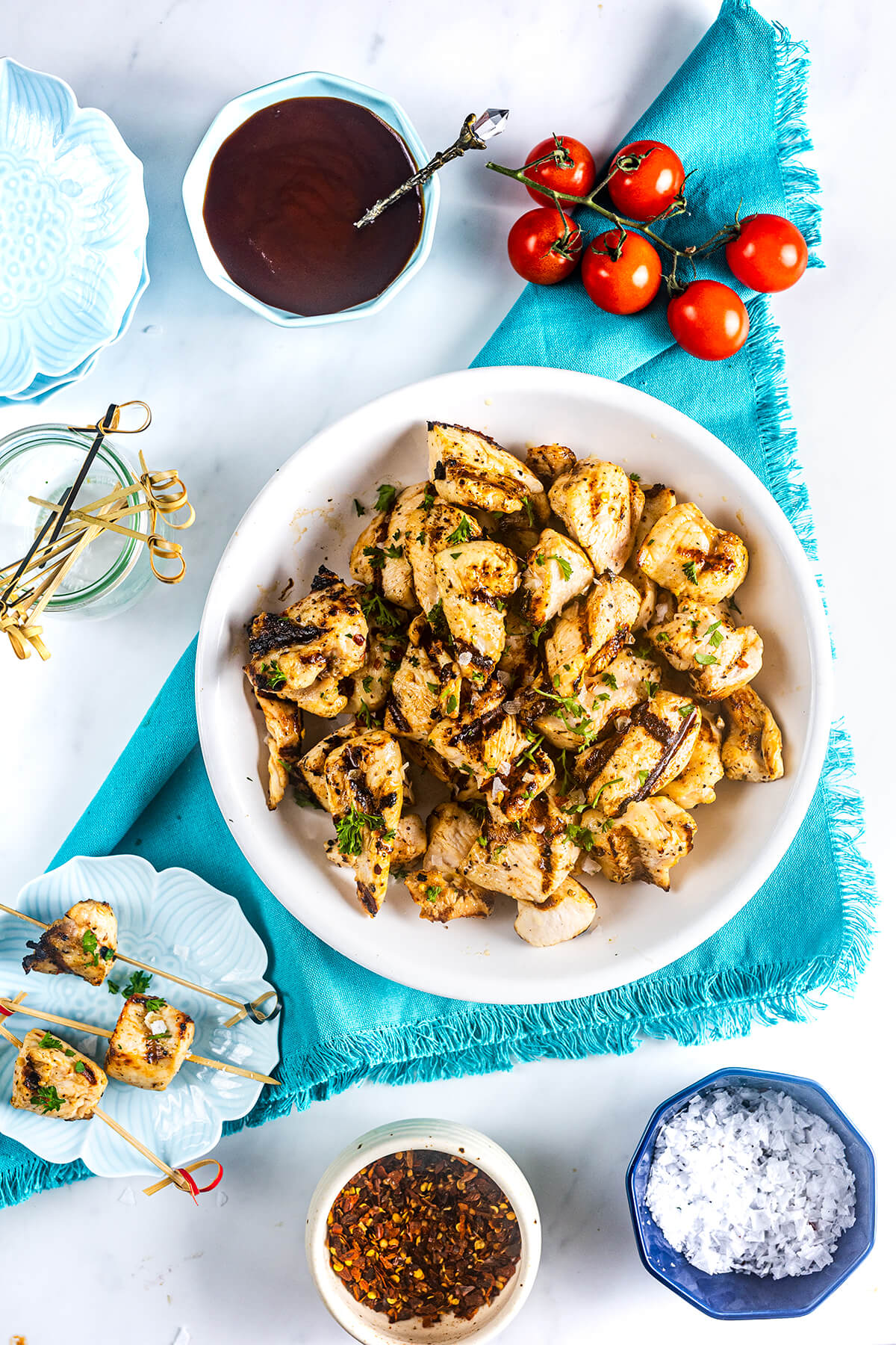 A white plate of grilled chicken skewers garnished with herbs sits on a blue napkin. Surrounding it are cherry tomatoes, barbecue sauce, seasoning, salt, and extra skewers, all on a white surface.
