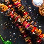Grilled beef and vegetable skewers with peppers, onions, and tomatoes on a dark stone surface, garnished with herbs, salt, chili flakes, and a sprig of parsley nearby.
