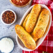 A basket of seasoned breadsticks on a red cloth, surrounded by bowls of marinara sauce, grated cheese, and red pepper flakes on a gray surface.